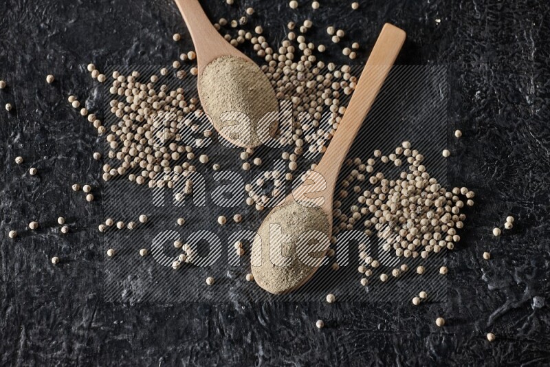 2 wooden spoons full of white pepper powder with white pepper beads on textured black flooring