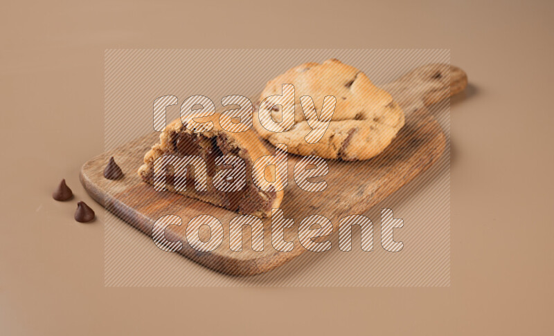 a chocolate chip cookie with another one cut in half on a wooden cutting board on a brown background