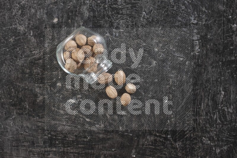 A glass spice jar full of whole nutmeg seeds flipped and the seeds spread out on a textured black flooring