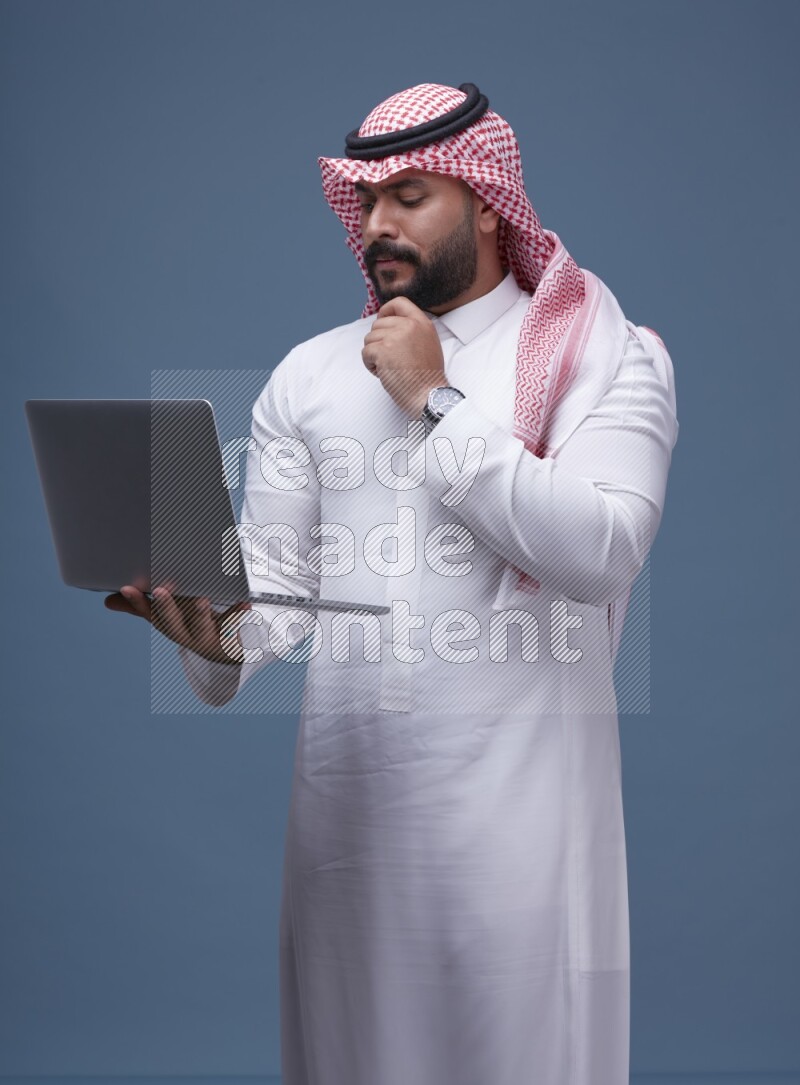 A man standing  with a laptop on Blue Background wearing Saudi Thob and Shomag