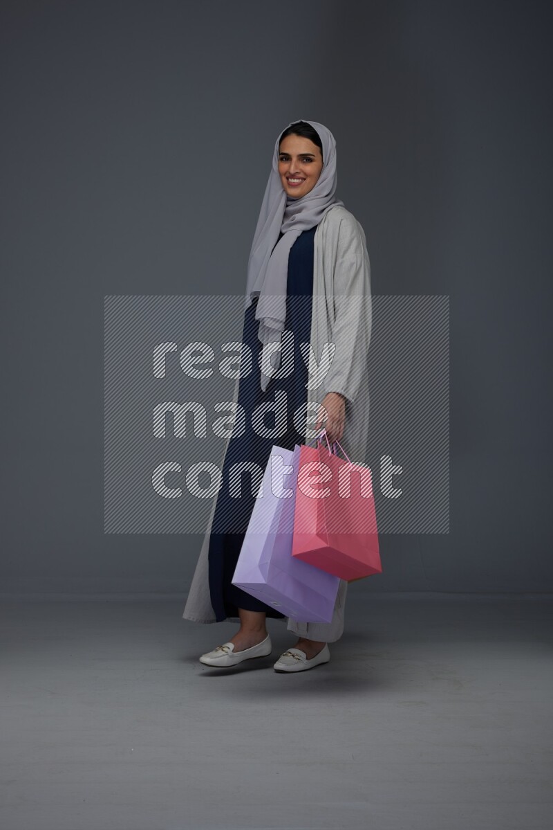 A Saudi woman wearing a light gray Abaya and head scarf standing and holding shopping bags on a grey background
