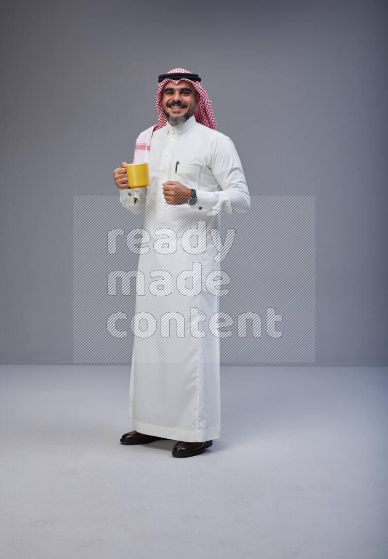 Saudi man Wearing Thob and red Shomag standing holding mug on Gray background