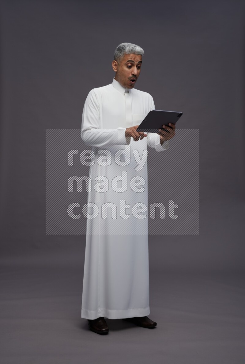 Saudi man wearing thob standing working on tablet on gray background