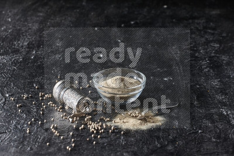 A glass bowl full of white pepper powder with pepper beads, a metal grinder and a metal spoon on textured black flooring