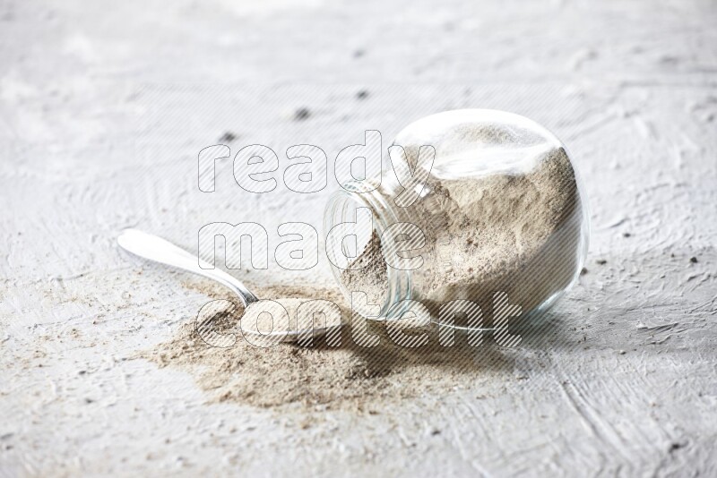A flipped herbal glass jar and metal spoon full of white pepper powder with spilled powder on textured white flooring