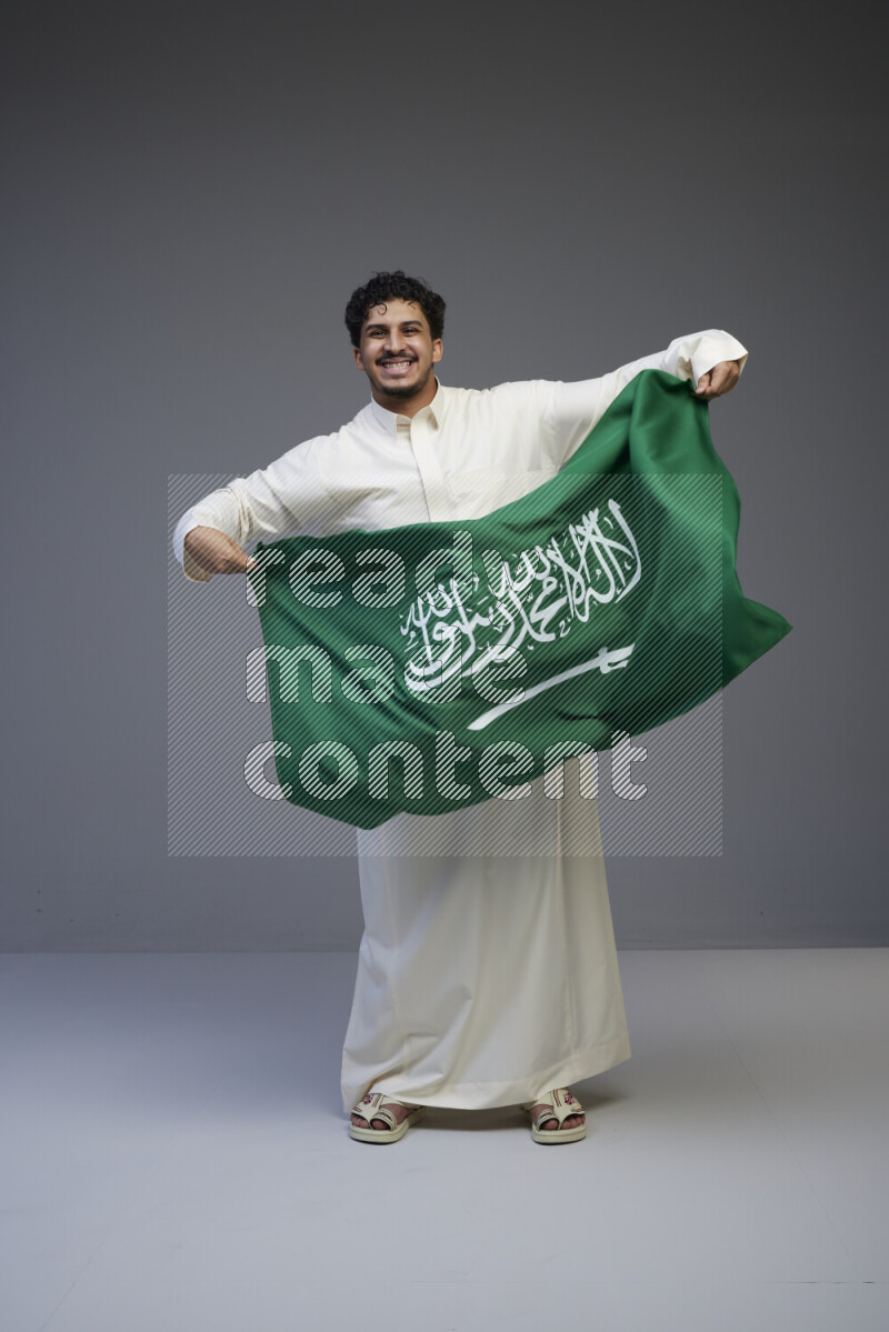 A saudi man standing wearing thob holding big saudi flag on gray background