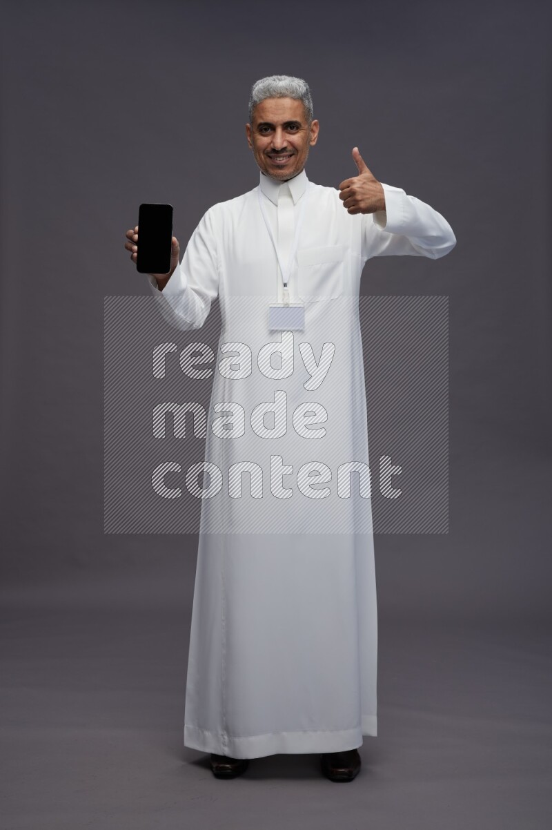 Saudi man wearing thob with neck strap employee badge standing showing phone to camera on gray background