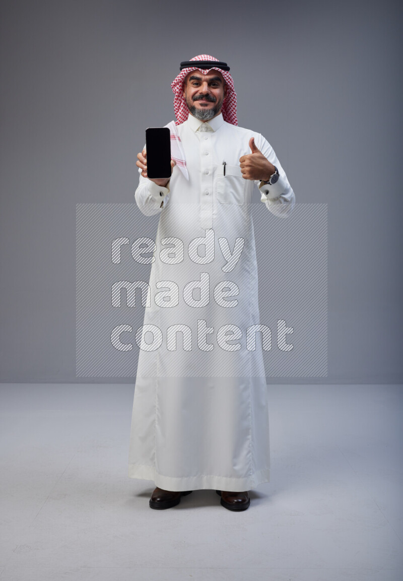 Saudi man Wearing Thob and red Shomag standing showing phone to camera on Gray background