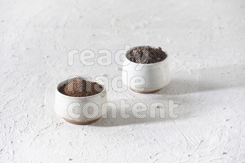 2 Beige ceramic bowls, one full of cloves and the other full of powder on textured white flooring