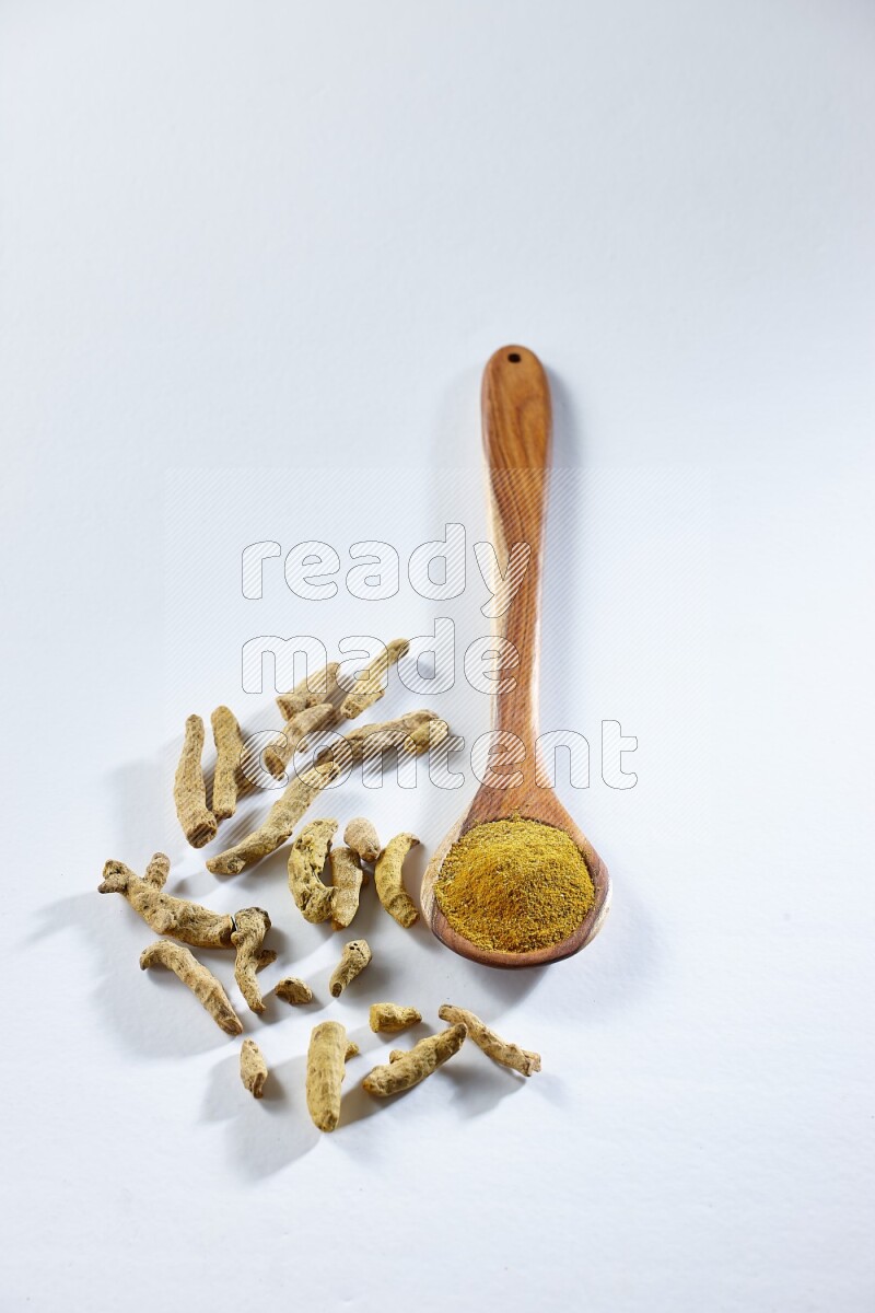 A wooden ladle full of turmeric powder and dried turmeric fingers beside it on white flooring