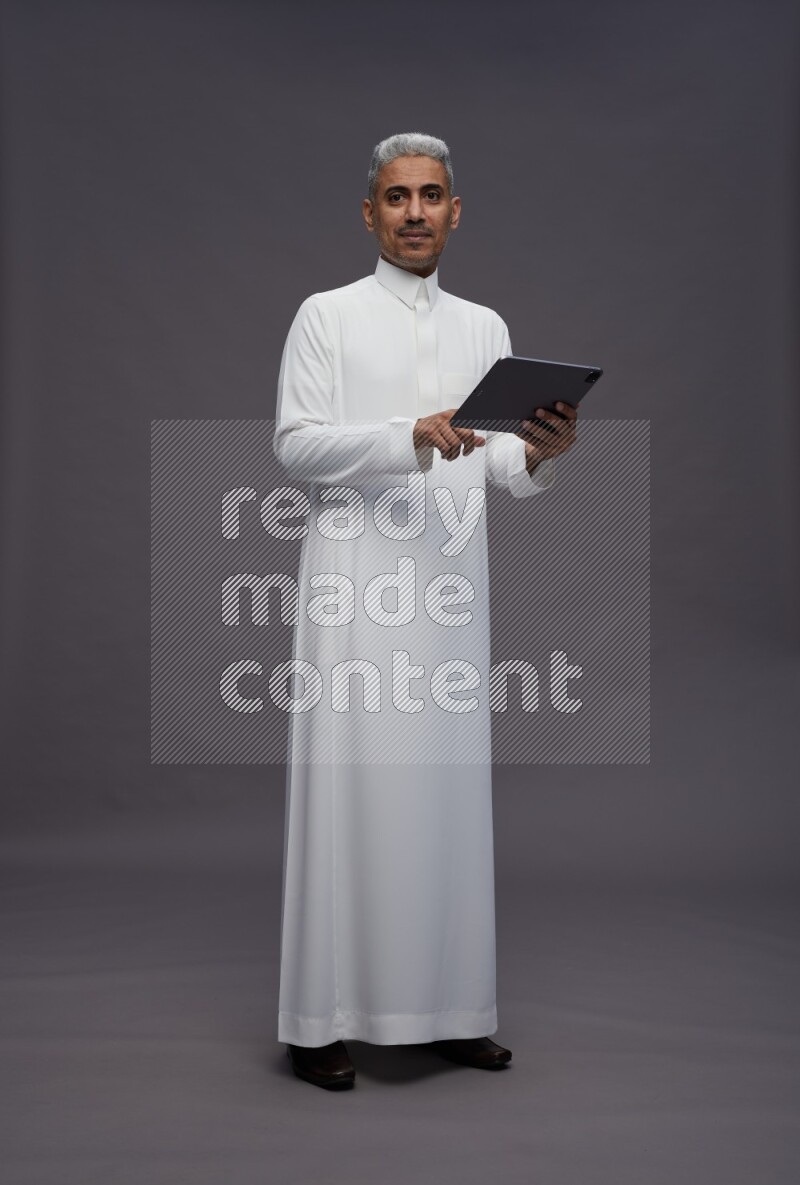 Saudi man wearing thob standing working on tablet on gray background