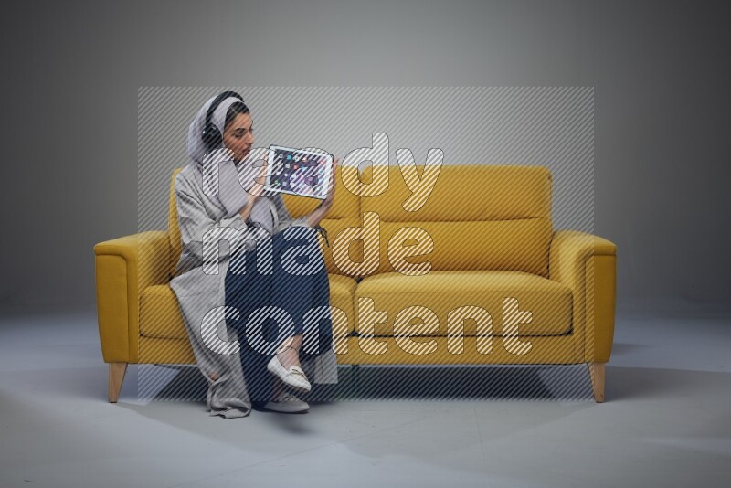 A Saudi female wearing a light gray Abaya and head scarf sitting on a yellow sofa and showing her tablet's screen while wearing headphones eye level on a grey background
