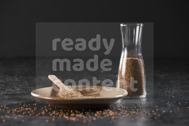 A beige pottery plate full of mustard seeds and a wooden spoon in it with a glass jar filled with the seeds on a textured black flooring