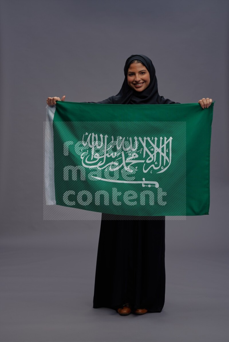 Saudi woman wearing Abaya standing holding Saudi flag on gray background