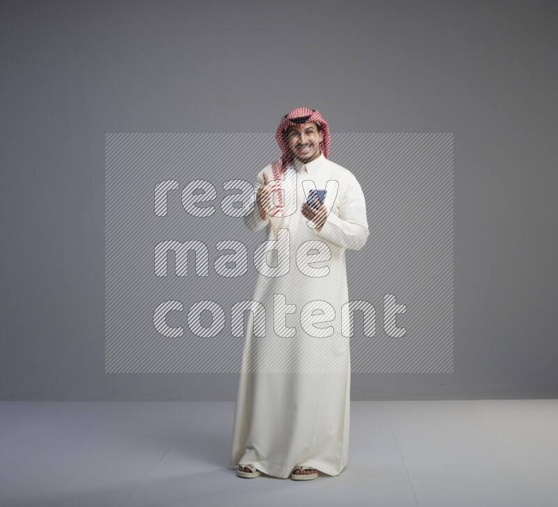 A  Saudi man standing wearing thob and red shomag texting on phone on gray background