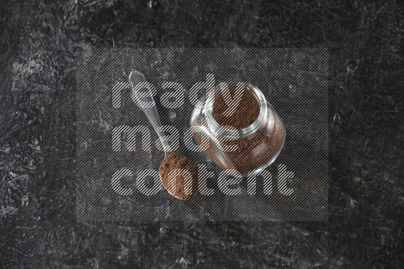A glass spice jar and a metal spoon full of cloves powder on textured black flooring