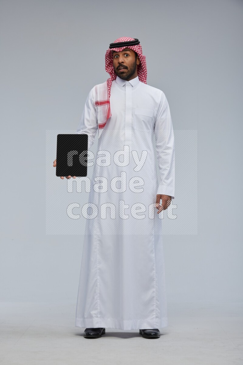 Saudi man Wearing Thob and shomag standing showing tablet to camera on Gray background