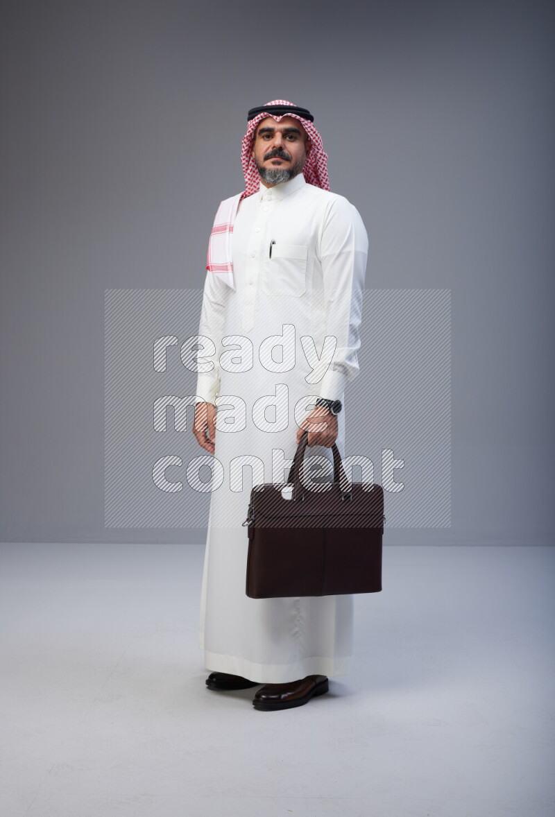 Saudi man Wearing Thob and red Shomag standing holding bag on Gray background