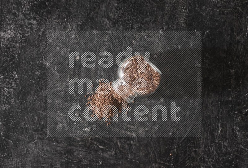 وعاء زجاجي مقلوب ممتلئ بحبوب بذر الكتان والحبوب متناثرة منه علي خلفية سوداء