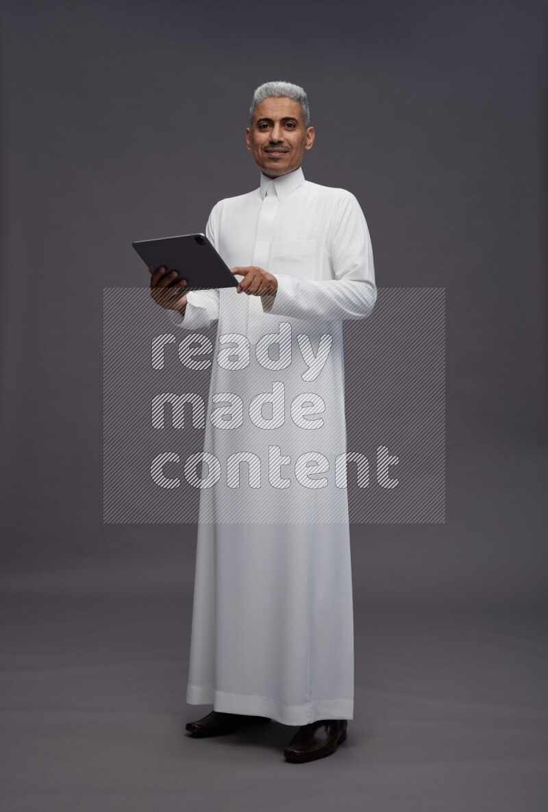 Saudi man wearing thob standing working on tablet on gray background