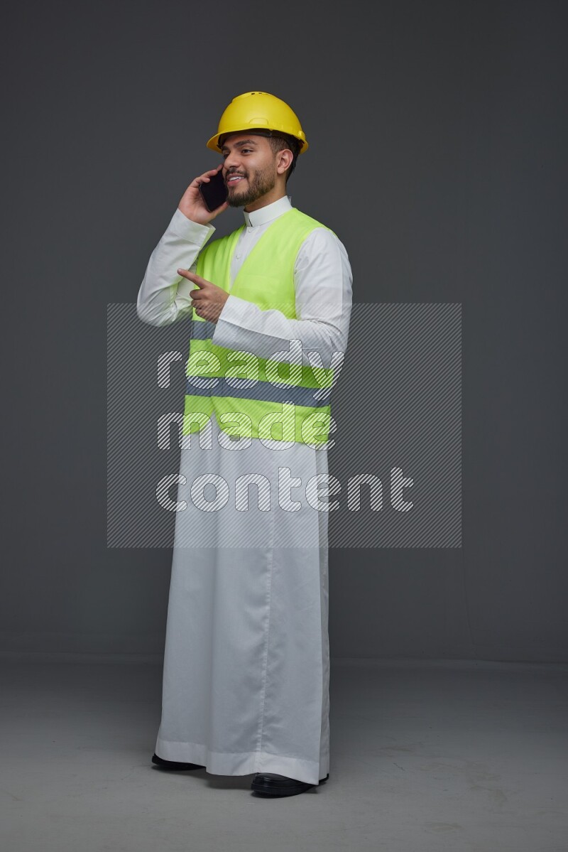 A Saudi man wearing Thobe with a yellow safety vest and white helmet standing and talking in the phone eye level on a gray background