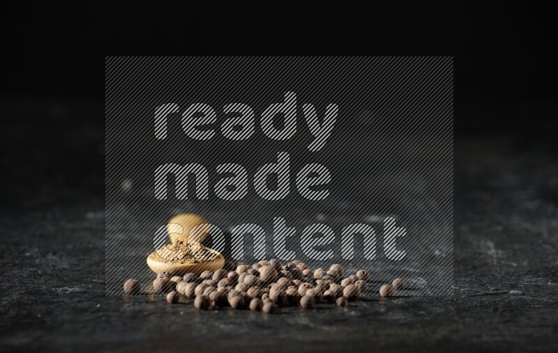 A wooden spoon full of allspice powder and whole balls spreaded on a textured black flooring