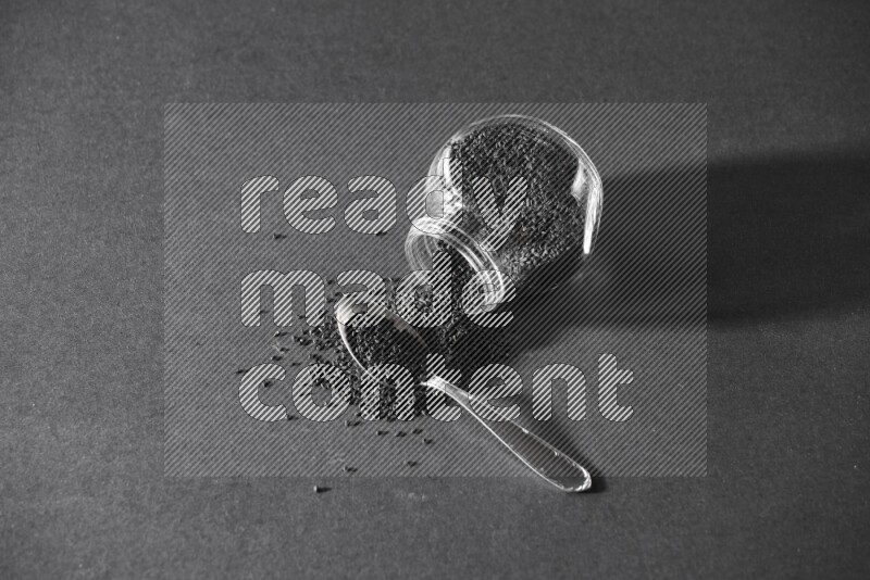 وعاء بهارات زجاجي مقلوب ممتلئ ببذور حبة البركة والبذور تناثرت منه مع ملعقة معدنية ممتلئة على أرضية سوداء