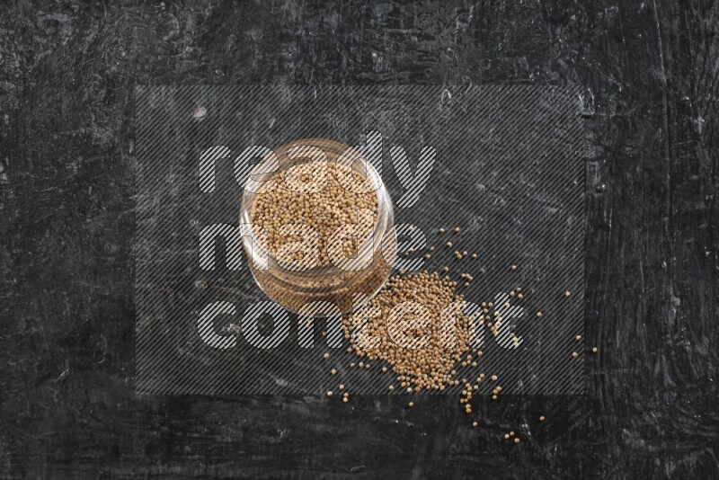 A glass jar full of mustard seeds and jar is flipped and seeds spread out on a textured black flooring