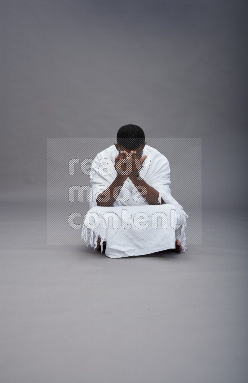 A man wearing Ehram sitting on floor dua'a on gray background