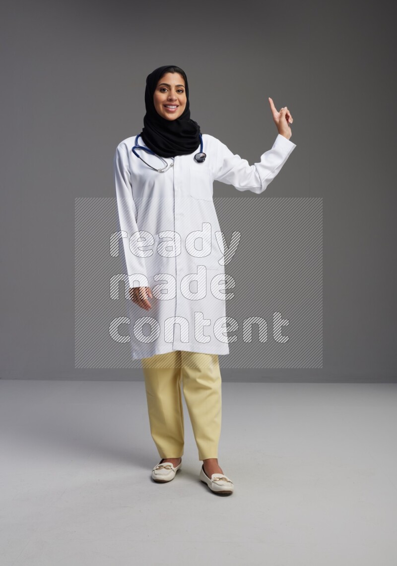 Saudi woman wearing lab coat with stethoscope standing interacting with the camera on Gray background