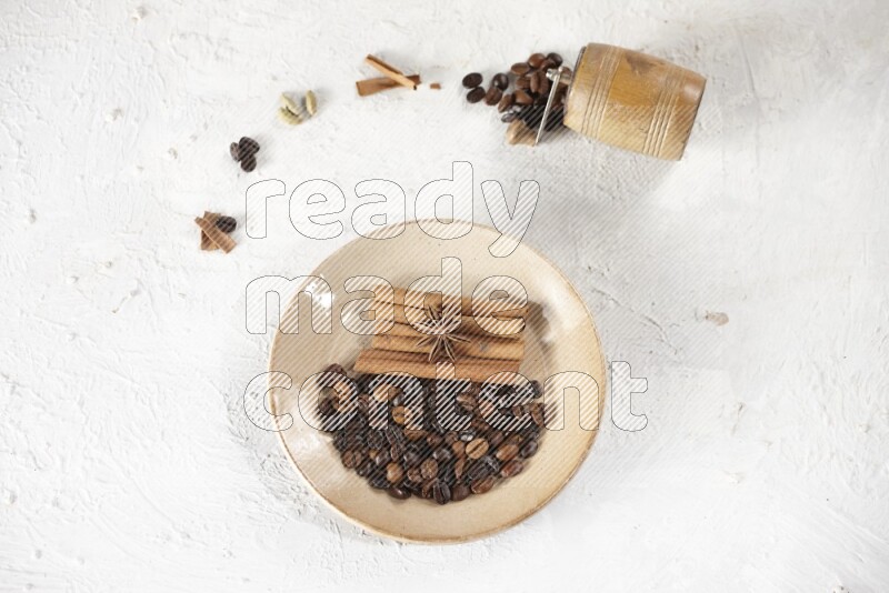 Beige plate full of coffee beans, cinnamon sticks and star anise with a coffee grinder, coffee beans, cinnamon pieces and cardamom next of it on white background