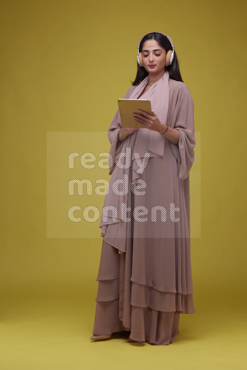 A woman Holding an Ipad with Headphone on a Yellow Background wearing Brown Abaya