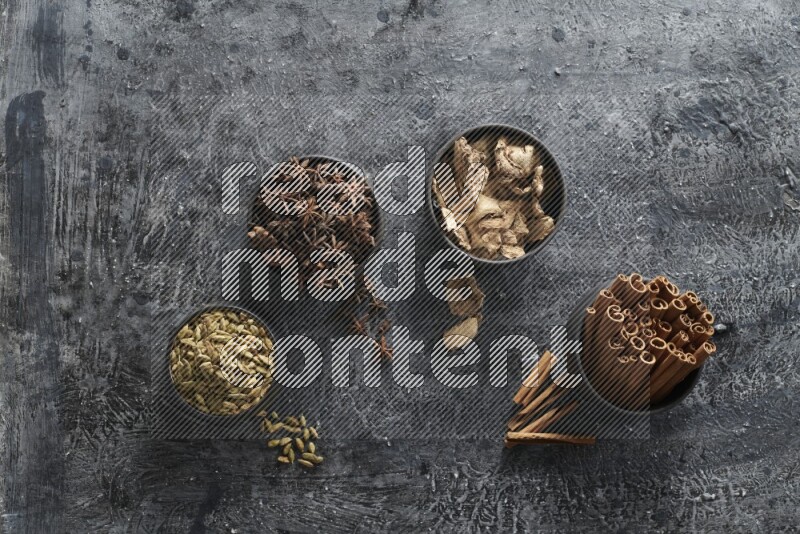 Ginger, Cardamom, Star anise and cinnamon sticks in 4 bowls on a textured black background