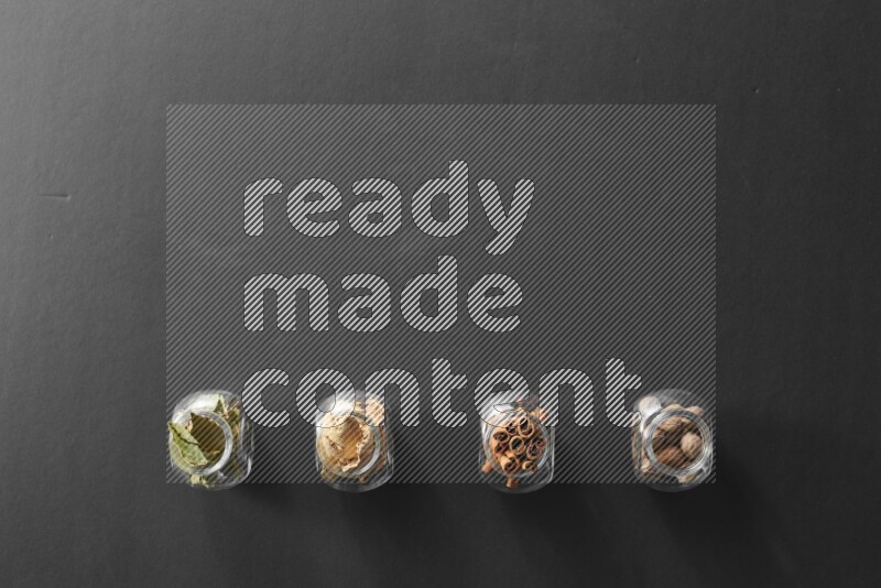 4 glass spice jars full of laurel bay leaves, ginger, cinnamon and nutmeg on black flooring