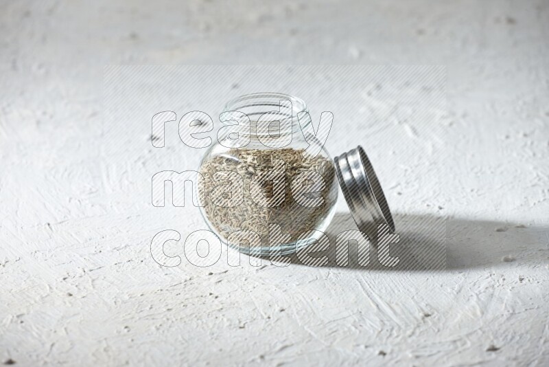 A glass spice jar full of cumin seeds on textured white flooring