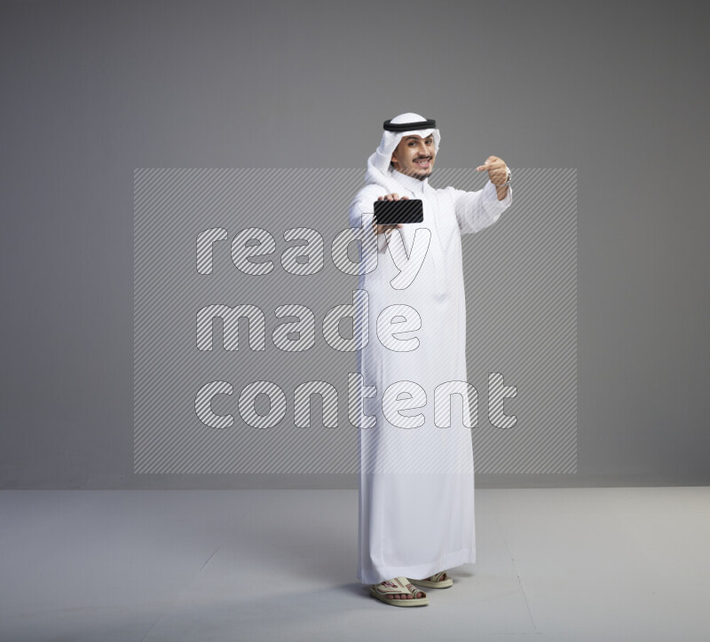 A Saudi man standing wearing thob and white shomag showing phone to camera on gray background