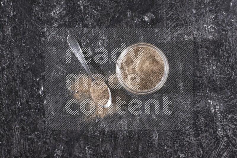 بودرة الفلفل الأسود في وعاء زجاجي مع ملعقة معدنية مليئة بالبودرة علي خلفية سوداء