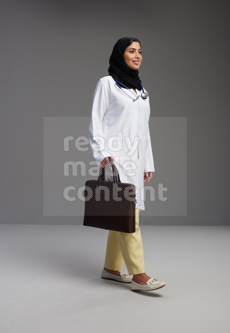 Saudi woman wearing lab coat with stethoscope standing holding bag on Gray background