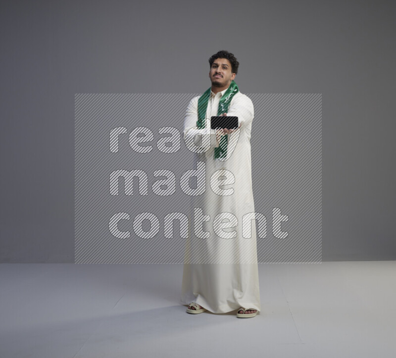A Saudi man standing wearing thob and Saudi flag scarf showing phone to camera on gray background