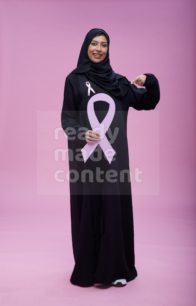 Saudi woman wearing pink ribbon on abaya standing holding awareness ribbon on plain pink background