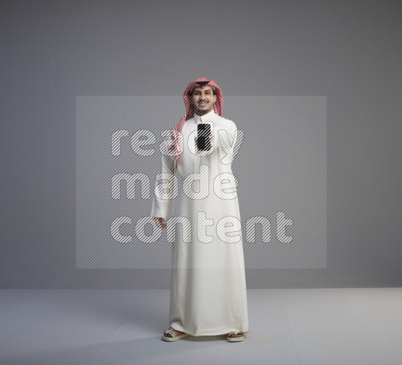 A Saudi man standing wearing thob and red shomag showing phone to camera on gray background