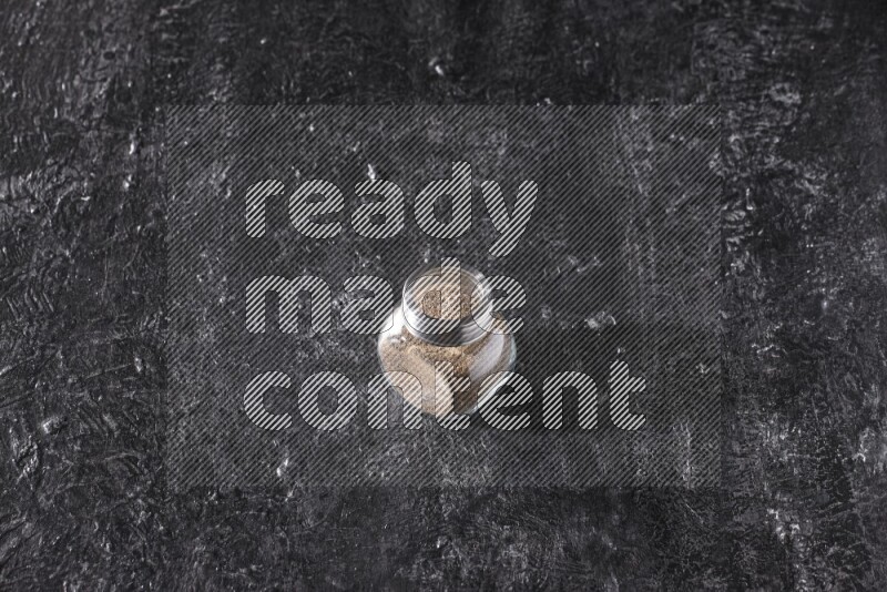 A glass spice jar full of black pepper powder on textured black flooring