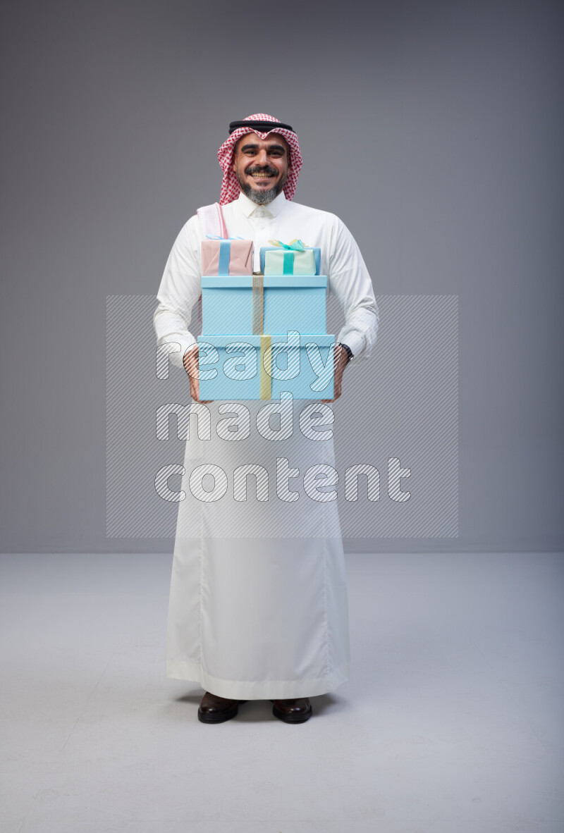 Saudi man Wearing Thob and red Shomag standing holding gift box on Gray background