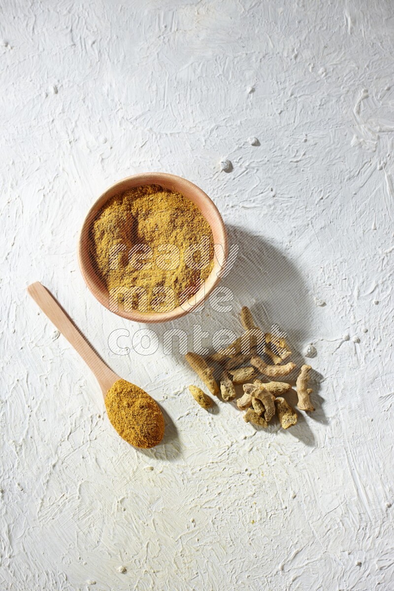 A wooden bowl and wooden spoon full of turmeric powder with dried turmeric fingers on textured white flooring