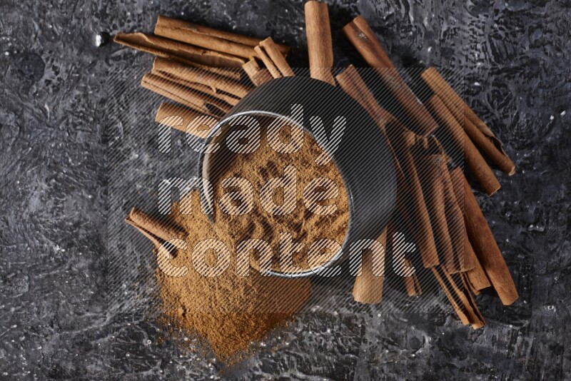 Black pottery bowl over filled with cinnamon powder and cinnamon sticks around the bowl on a textured black background