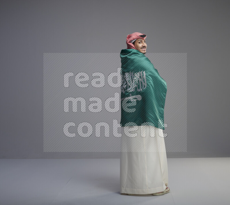 A Saudi man standing wearing thob and red shomag wrapping big saudi flag on gray background