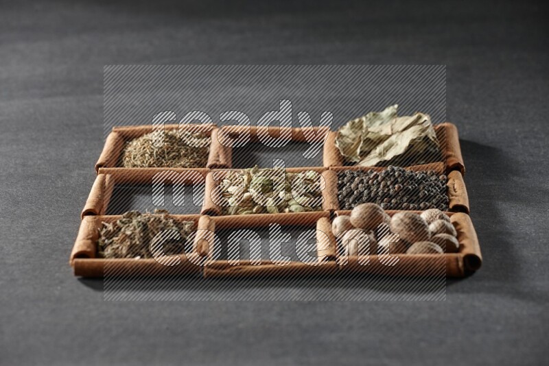 9 squares of cinnamon sticks full of cardamom in the middle surrounded by nutmeg, black pepper, bay laurel leaves, allspice, cumin, white pepper, dried basil and salt on black flooring