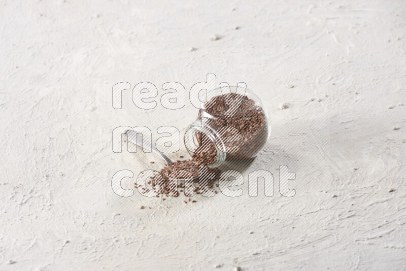A glass spice jar full of flax seeds flipped with a metal spoon full of the seeds on a textured white flooring