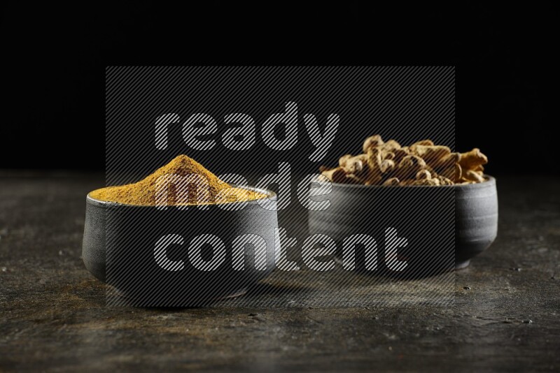 2 pottery black bowls, one full of turmeric powder and the other full of dried turmeric whole fingers on a textured black flooring
