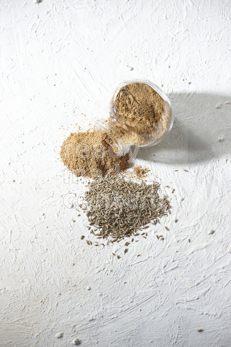 A flipped glass spice jar full of cumin powder and powder spilled out with cumin seeds on textured white flooring